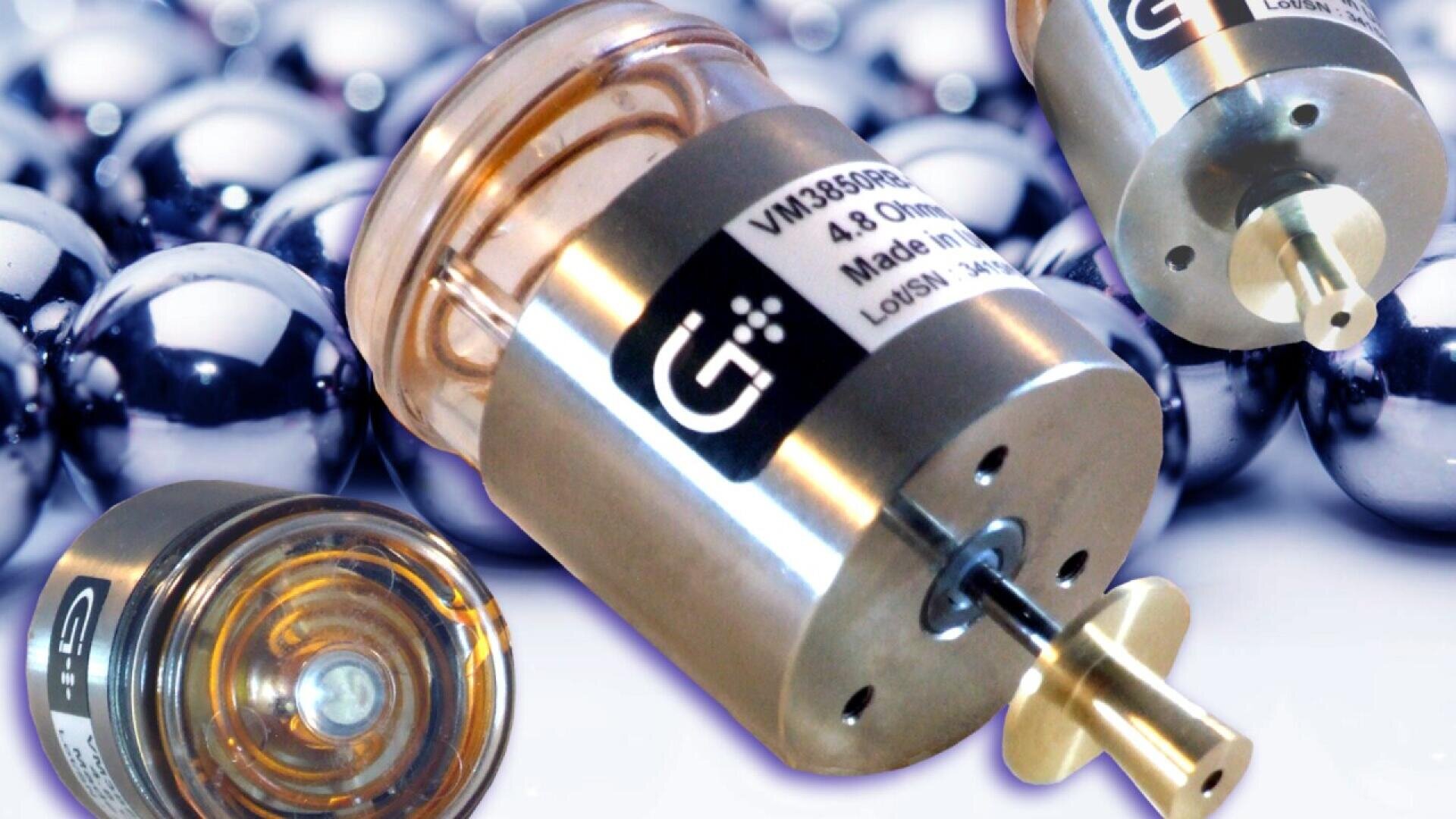 Close-up of several small metal cylindrical devices with labels, some showing a transparent end and a small shaft, placed against a background of shiny metallic spheres.
